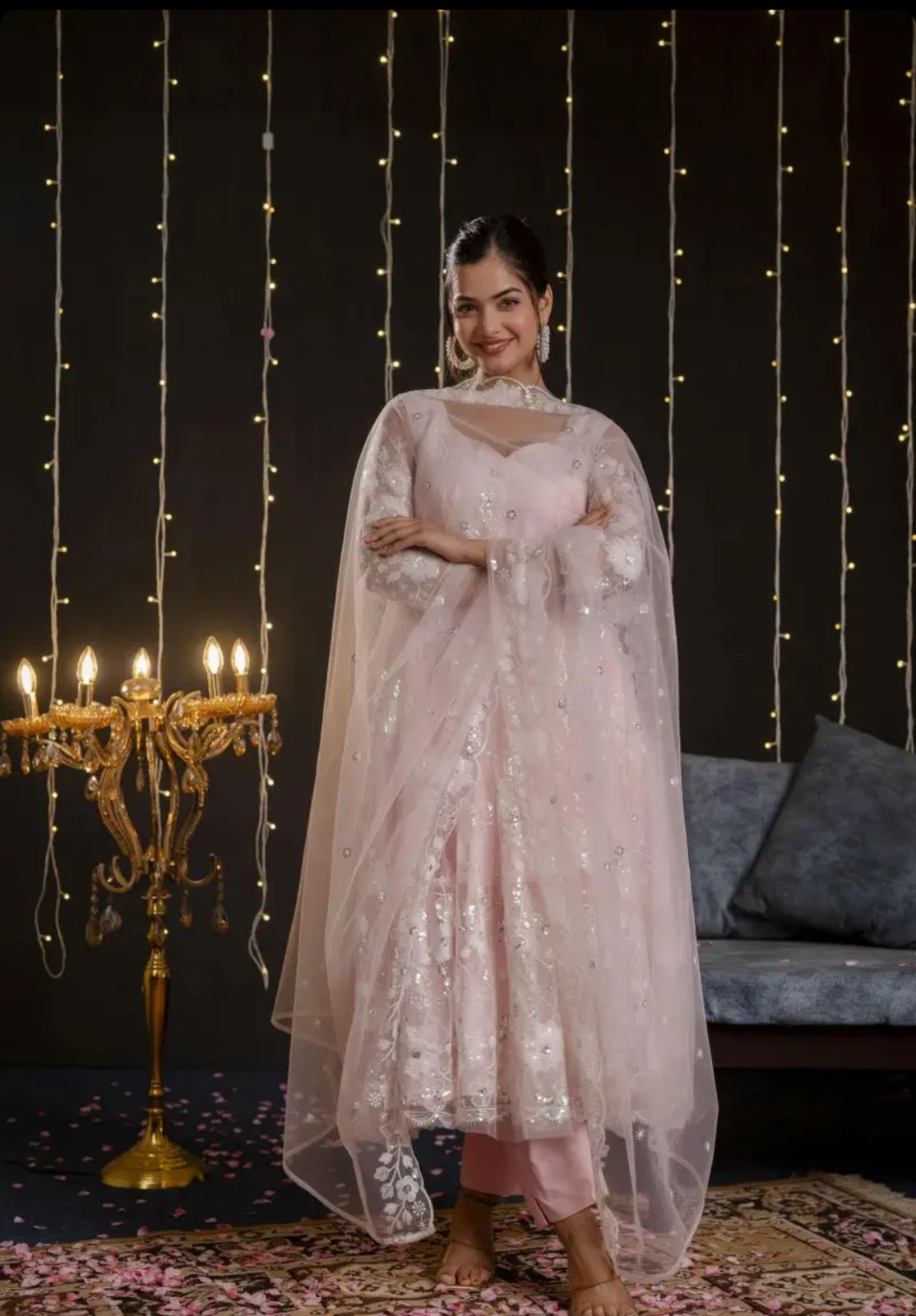 Woman in a light pink traditional outfit standing in a decorated room with string lights and a candelabra.