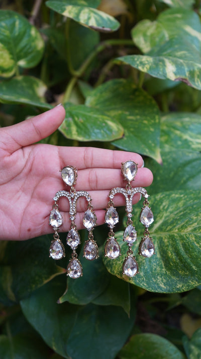 Silver-toned chandelier earrings with cascading teardrop crystals, hand-held against green leaves background – Noor Jharna by Pahraav.